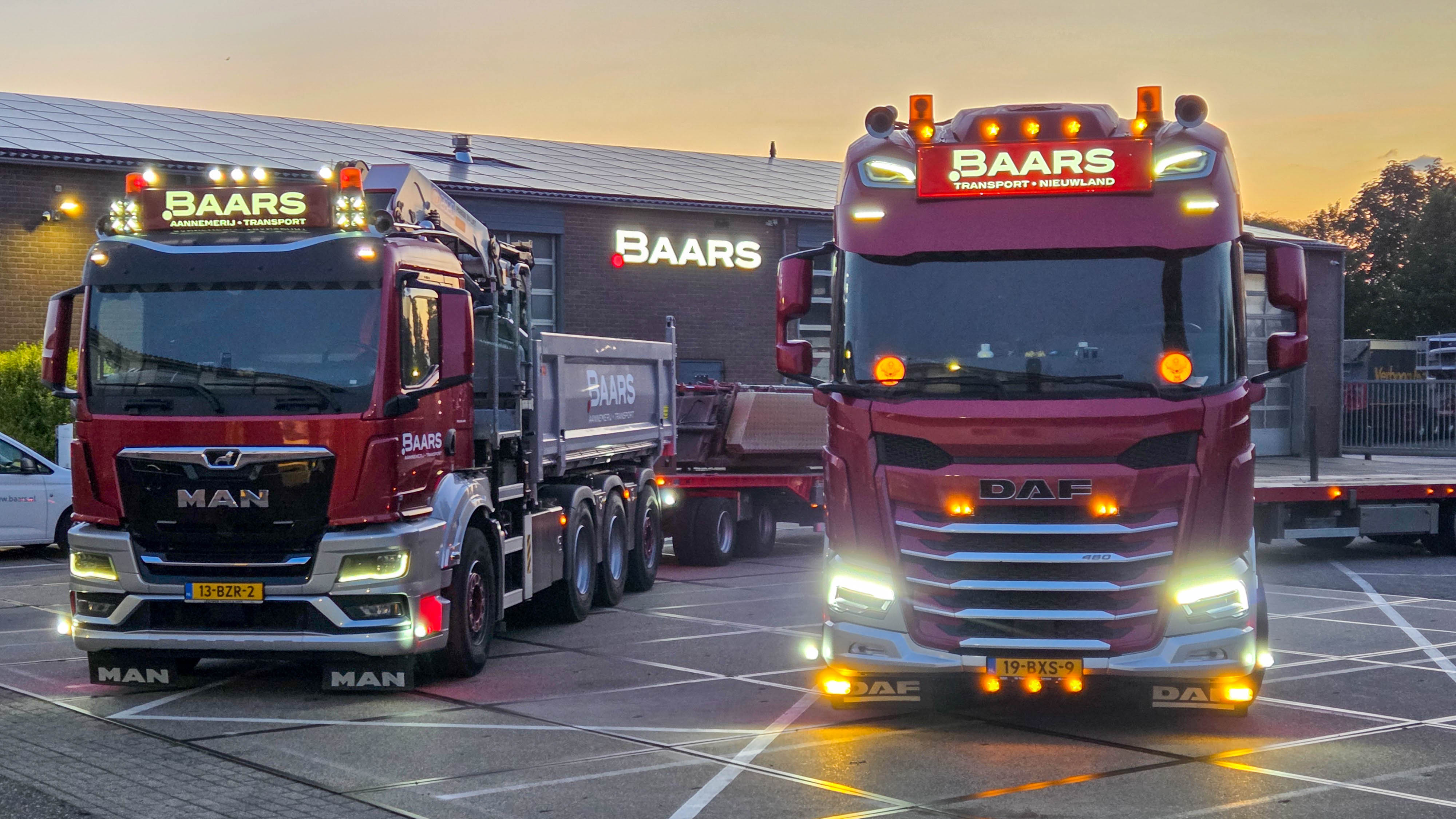 Twee vrachtwagens van Baars, een MAN en een DAF, geparkeerd voor het bedrijfspand bij schemering met verlichting aan.