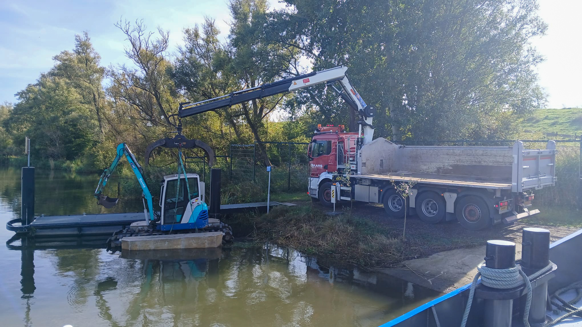 Een vrachtwagen van Baars met laadkraan tilt een kleine graafmachine op een ponton in het water, voor baggerwerkzaamheden in de Biesbosch.