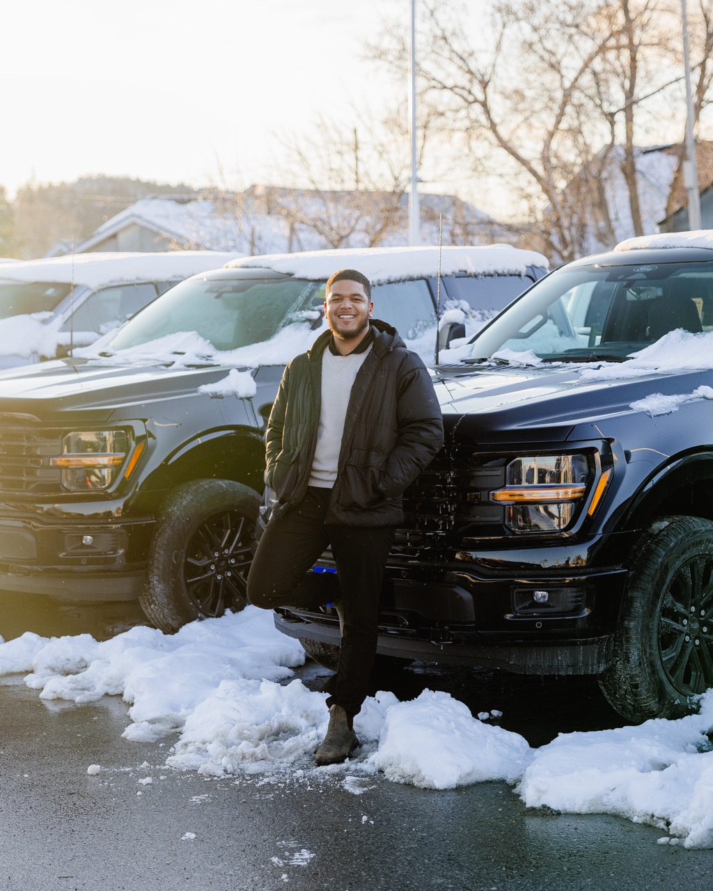 Smiling man in a black jacket leaning on a black pickup truck covered with snow in a snowy parking lot during daytime.