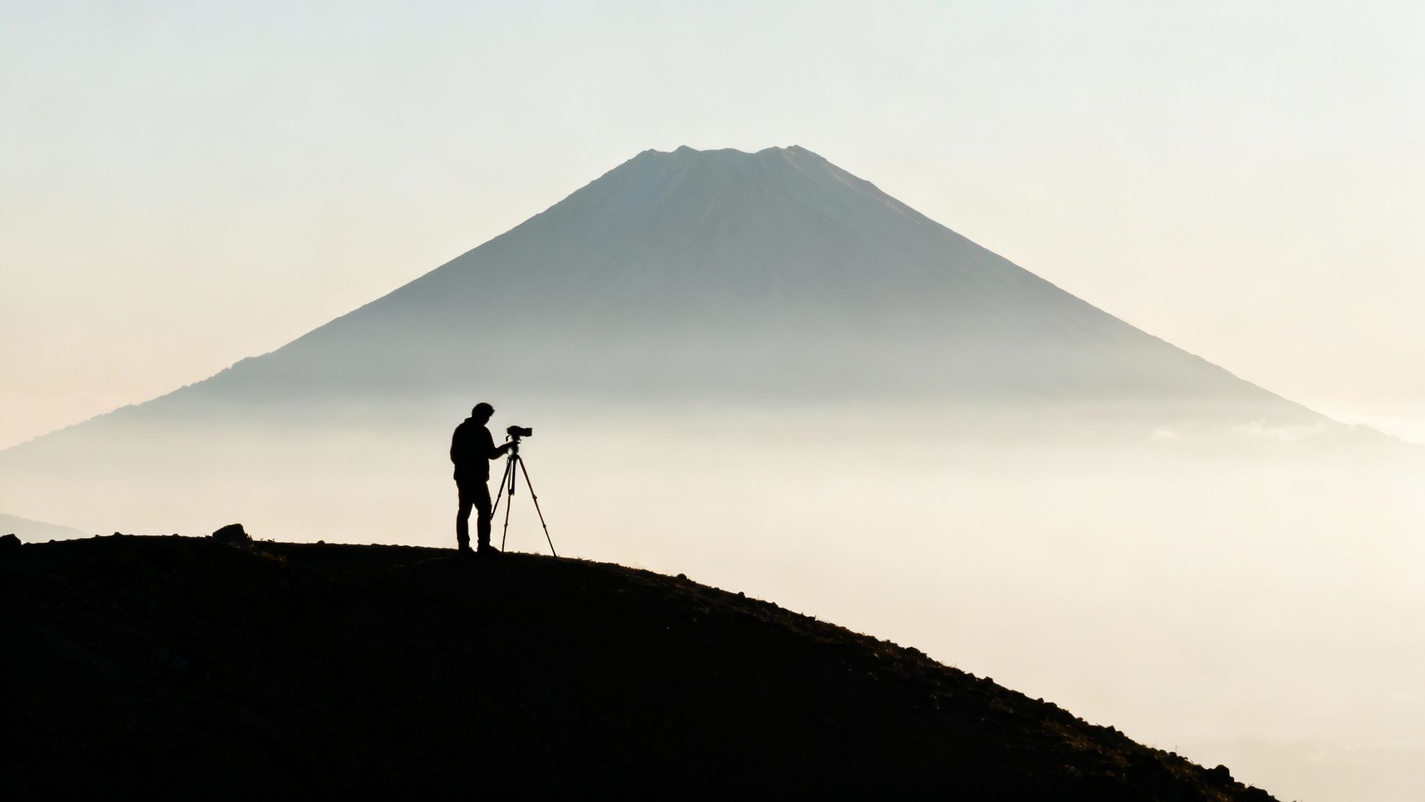 Silhouette of a photographer capturing a misty mountain landscape at sunrise with a camera and tripod.