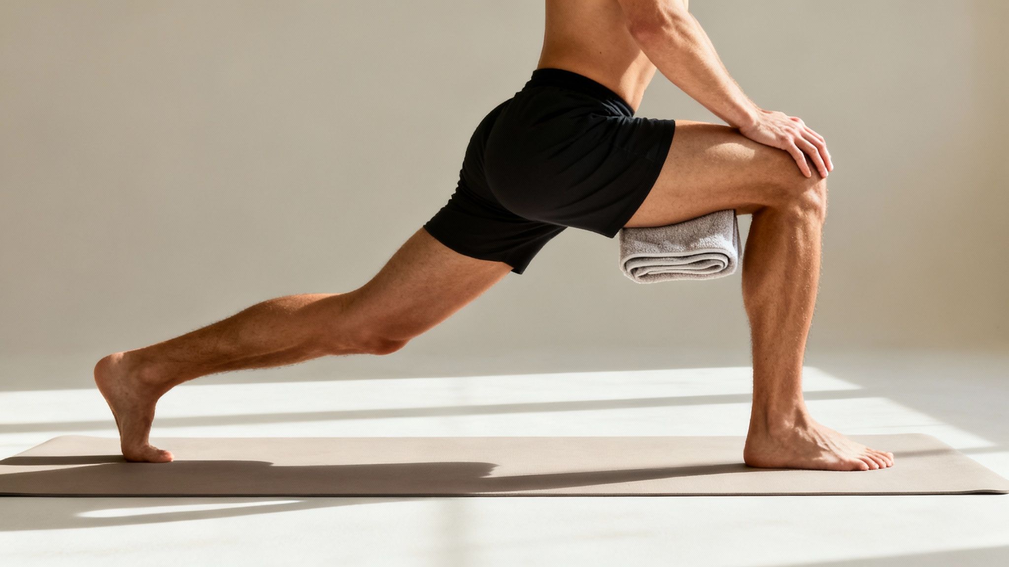 A man performs a static lunge stretch on a yoga mat with a towel under his front knee.