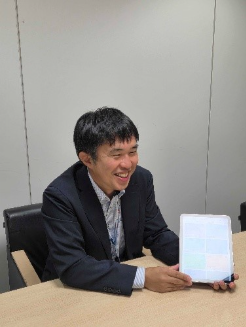 The image shows a person dressed in a suit, sitting at a desk with a tablet device displaying a colorful, grid-like interface.AI 生成コンテンツは誤りを含む可能性があります。