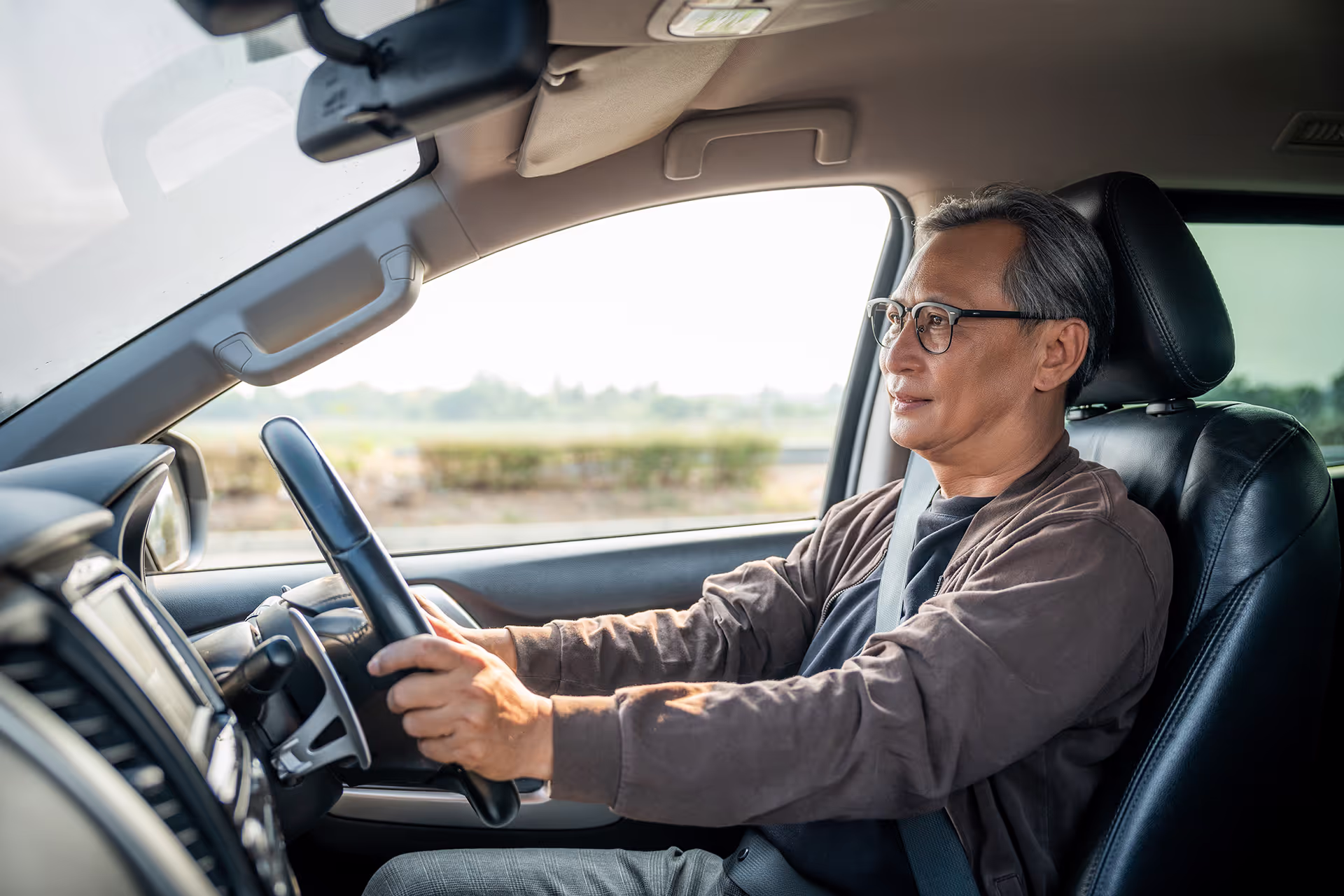 A middle-aged man wearing glasses and a seatbelt drives a car, looking calm and focused, with a scenic outdoor view visible through the window.
