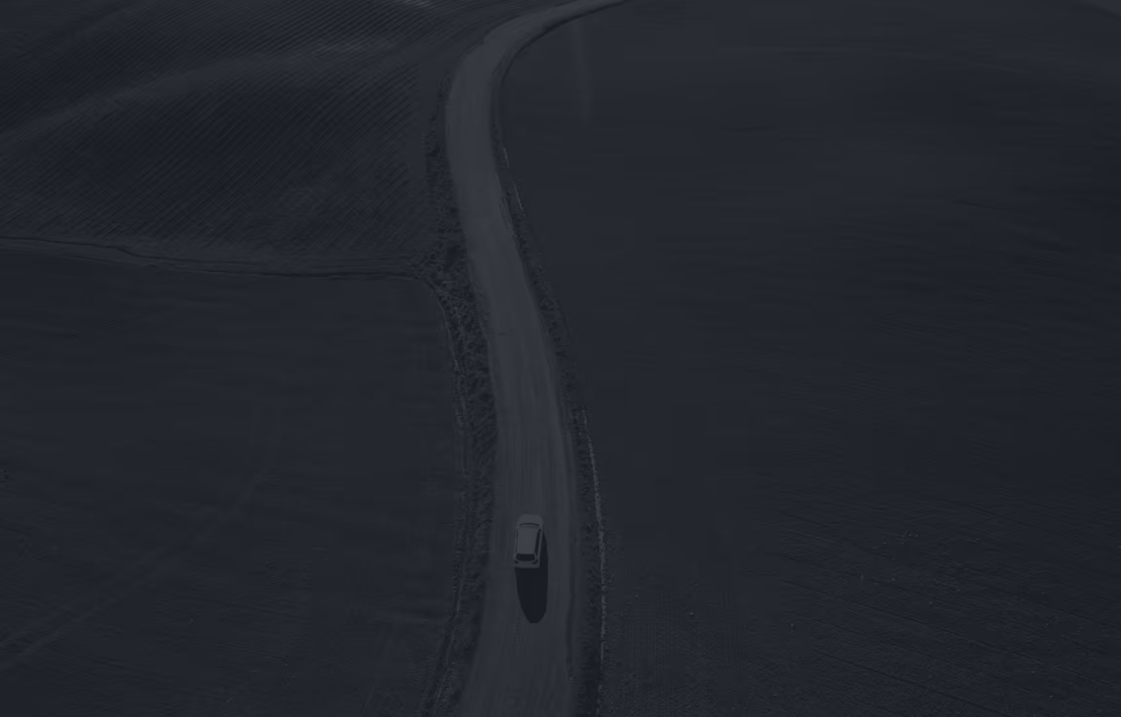 A lone car driving on a narrow winding road through dark, empty fields at dusk.