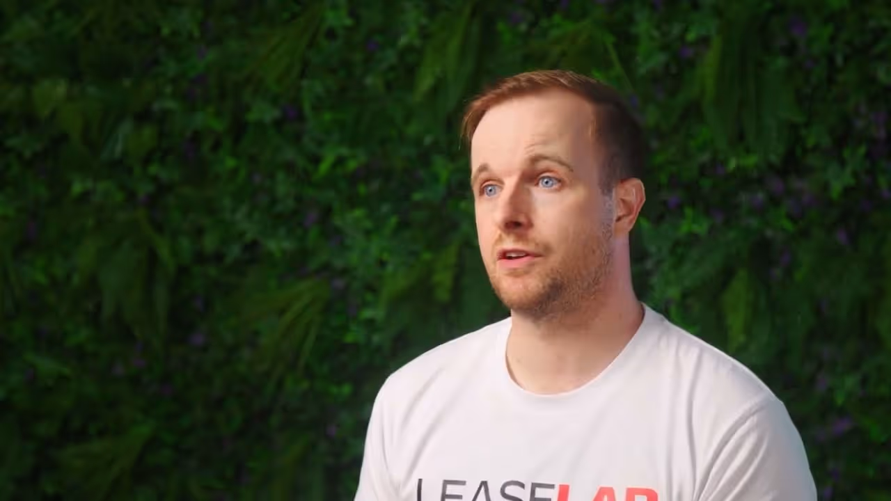 Man with light brown hair and blue eyes wearing a white LeaseLab t-shirt against a green leafy background.