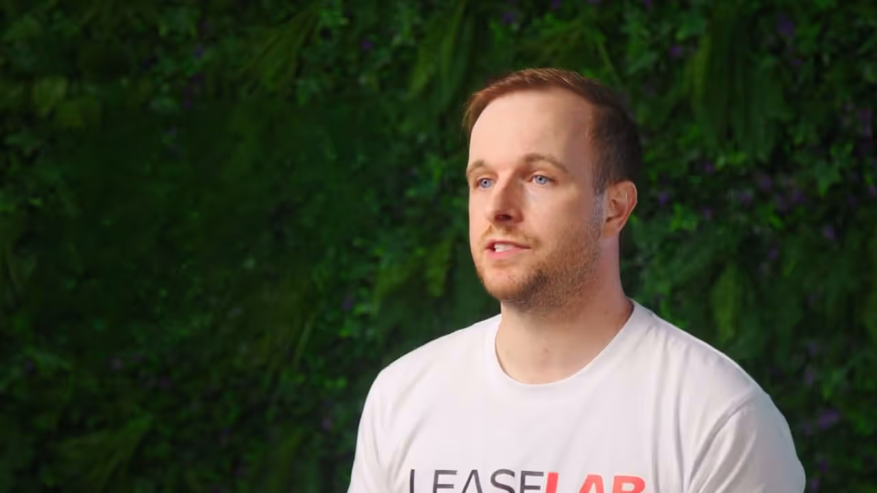 Man with light brown hair and blue eyes wearing a white LeaseLab T-shirt against a green leafy background.