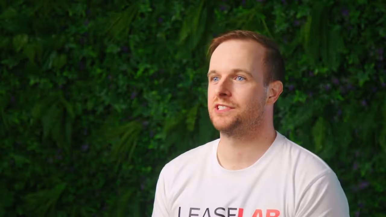 Man with short hair and blue eyes wearing a white LEASELAB T-shirt in front of a green leafy background.