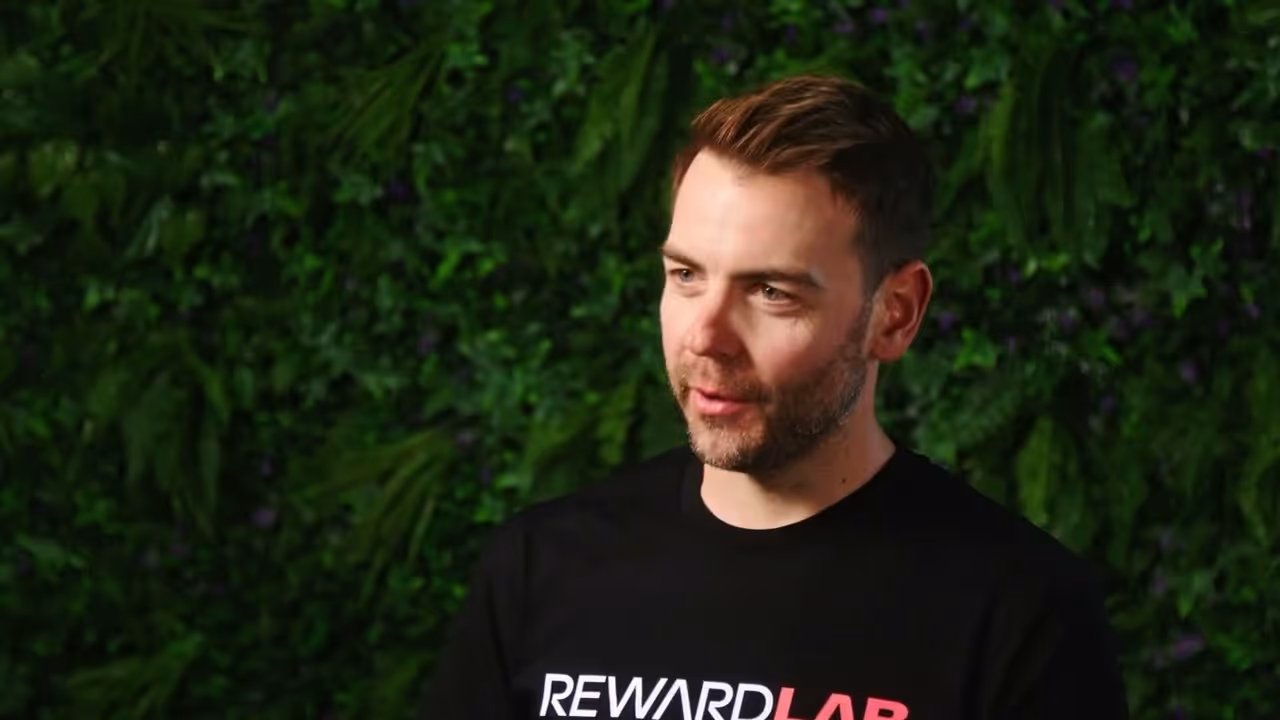 Man with short brown hair and beard wearing a black RewardLab T-shirt in front of a green foliage background.
