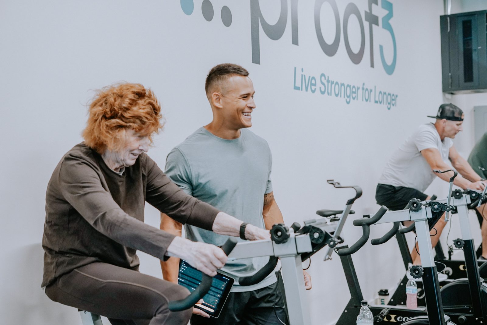 A proof3 coach smiles beside a member riding an exercise bike during a small-group session.