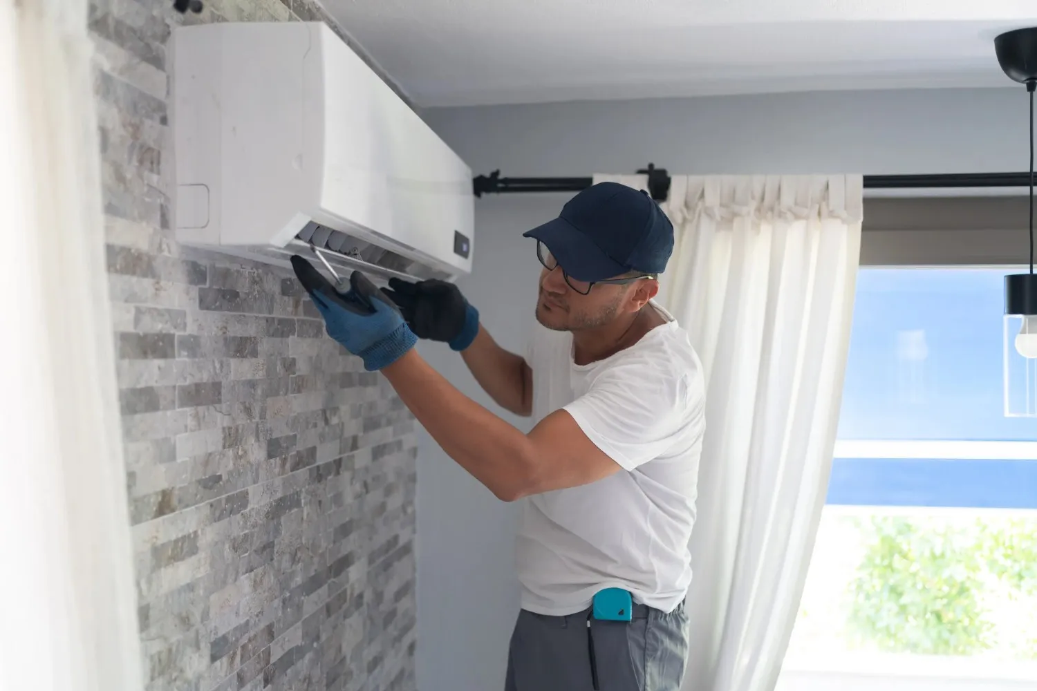 A person wearing white tee and blue cap is repairing a Ductless AC unit