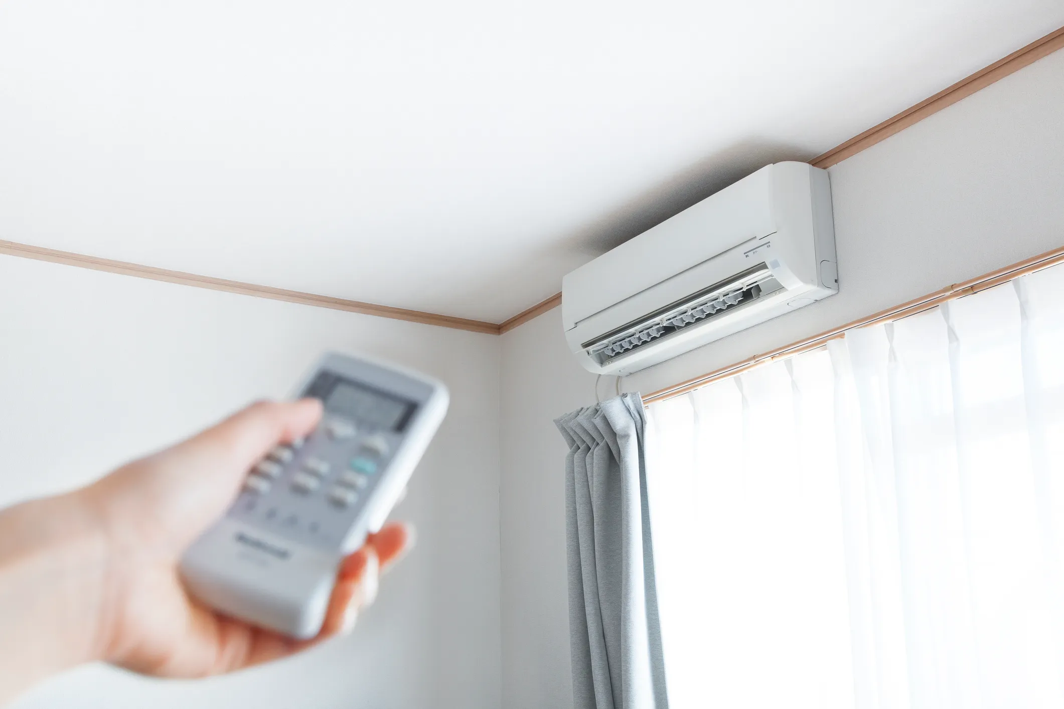 Person using remote to operate wall-mounted air conditioner in bright room with white walls and window