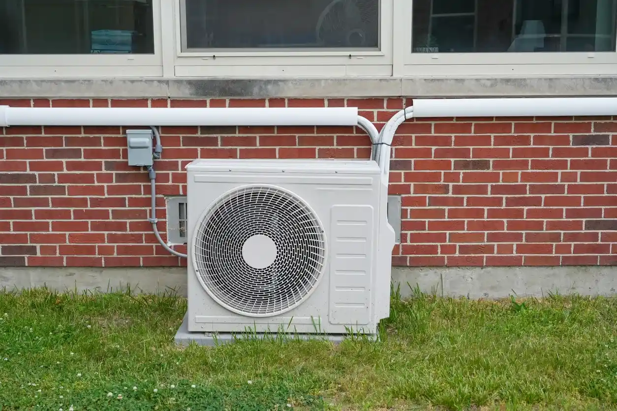 Outdoor air conditioning unit against brick wall with wiring, piping, and grass foreground