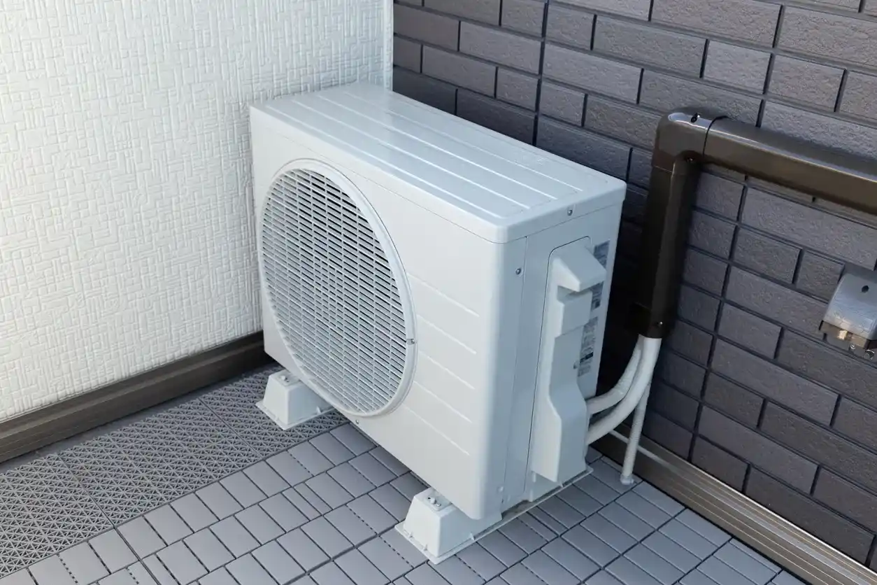 Outdoor air conditioning unit on tiled floor beside white-gray brick wall with connected pipes