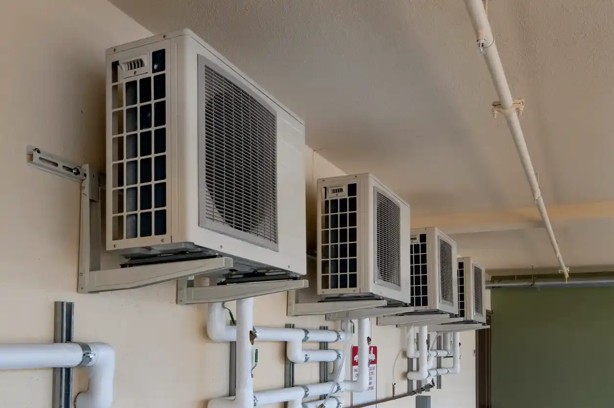 Row of four wall-mounted outdoor air conditioning units with protective grilles and exposed ceiling pipes