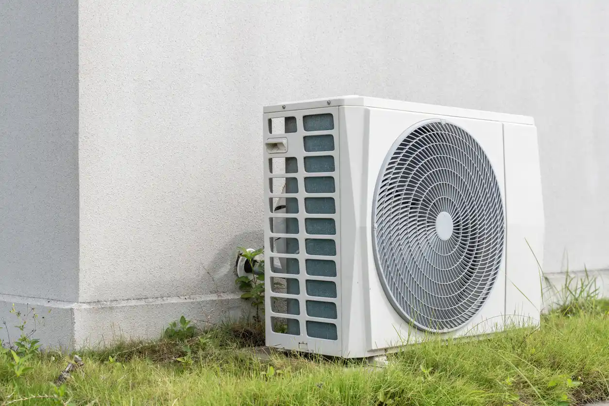 Outdoor air conditioning unit on ground beside building wall with fan, grilles, and nearby grass