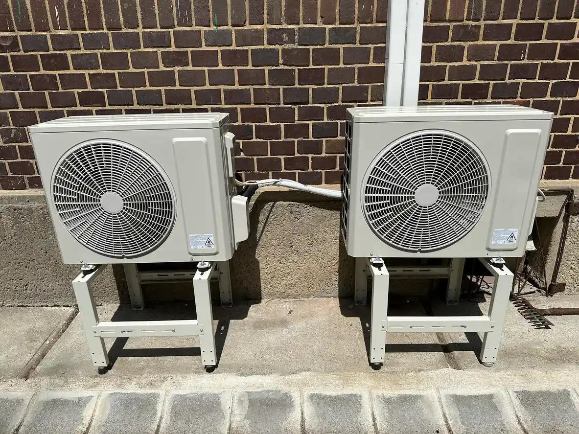 Two outdoor air conditioning units on metal stands against brick wall with connecting pipes and cables