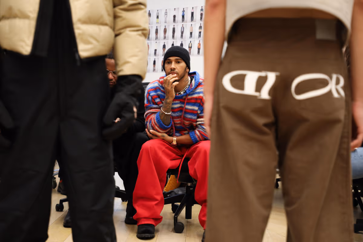 A man in a red and blue striped sweater sits in a chair while wearing a black beanie. He is looking at something in the distance. There is a person in brown pants with the word "DIOR" embroidered on the back standing in front of him. The person is blocking part of the man's view.