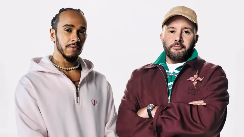 A portrait of Lewis Hamilton and a man wearing a brown baseball cap, standing next to each other with serious expressions on their faces. Hamilton is wearing a pink hoodie and a pearl necklace, while the other man is wearing a maroon jacket. Both men are looking directly at the camera.