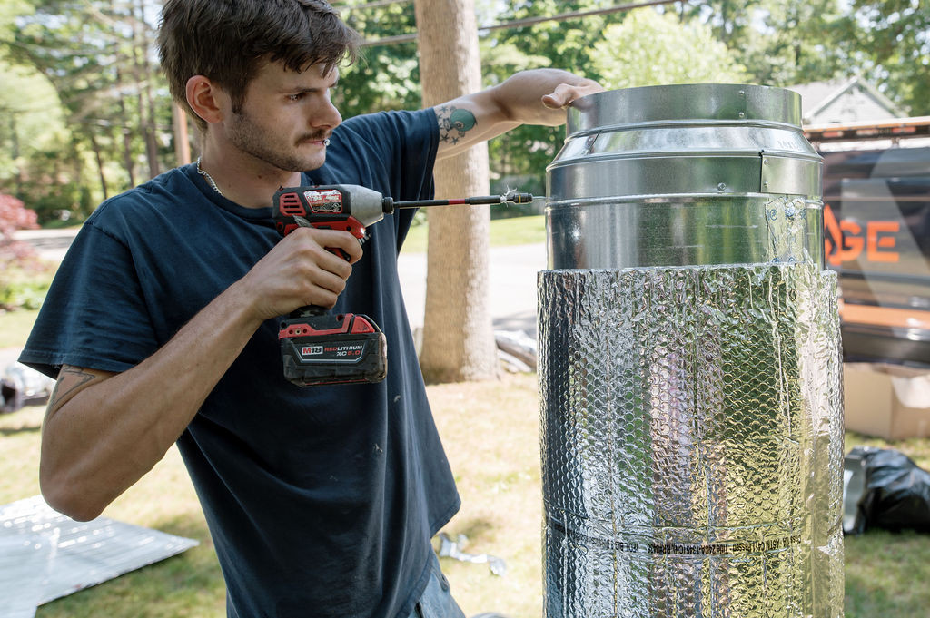 A Forge installer drilling into a duct trunk on the lawn beside the company van