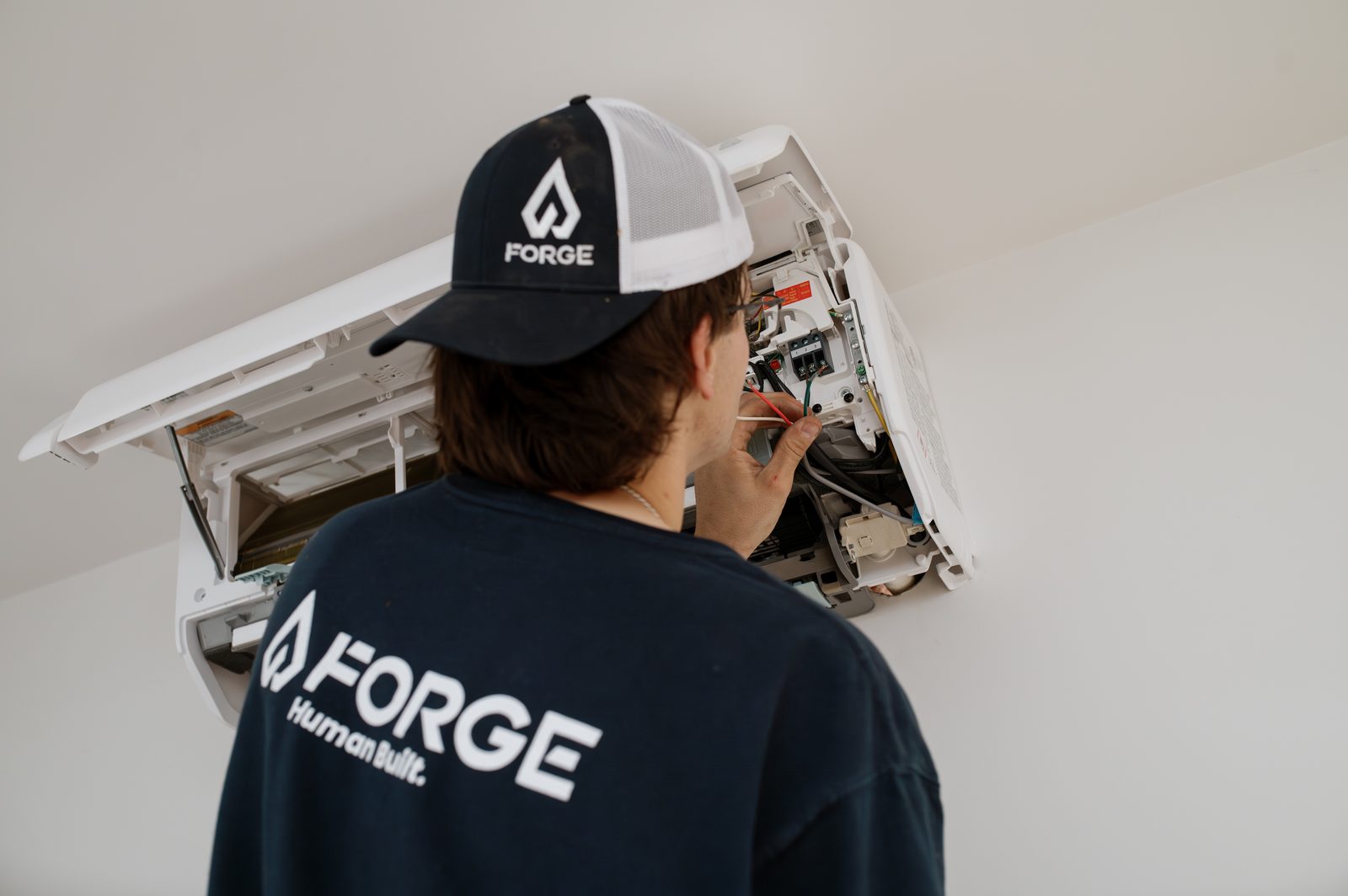 A Forge installer wiring an indoor mini split head, Forge Human Built on the back of his shirt