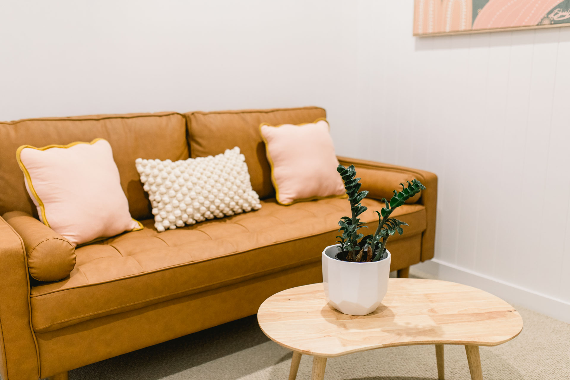 A bright room with a leathered sofa with decorative pillows, a table with a green plant, and colored painting on the wall.