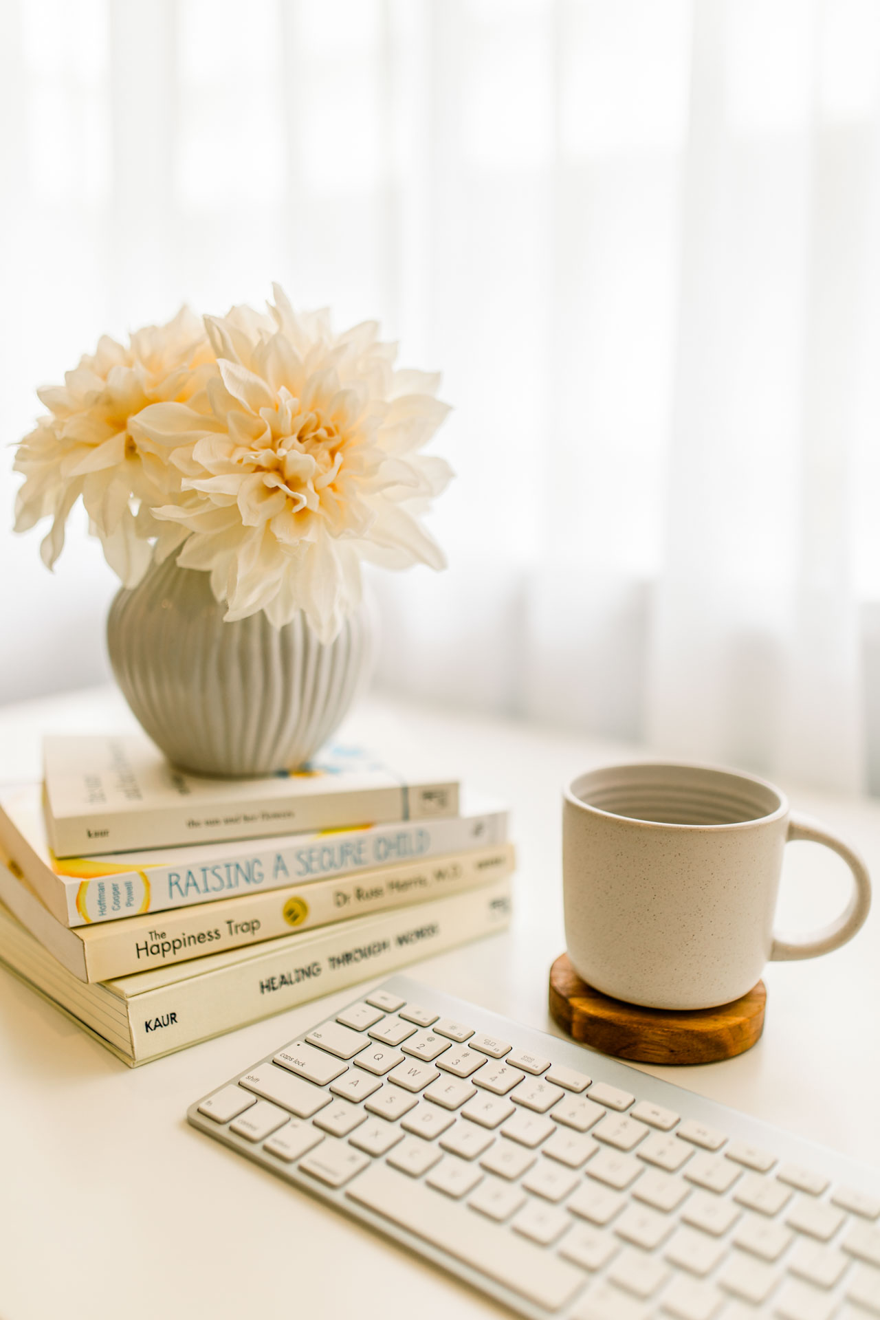 A well-organised modern work desk with a pile of books with an elegant flower vase, a cup of coffee, a laptop keyboard in a white elegant surroundings.
