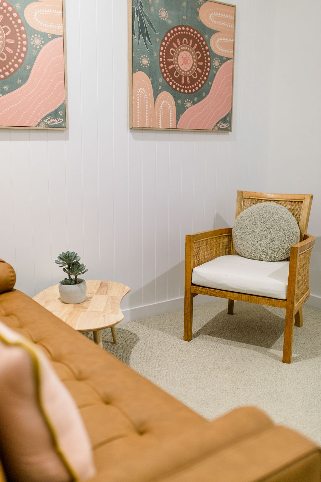 A cozy room with a chair, a green plant, a brown leathered sofan and a vibrant painting hanging on the wall.