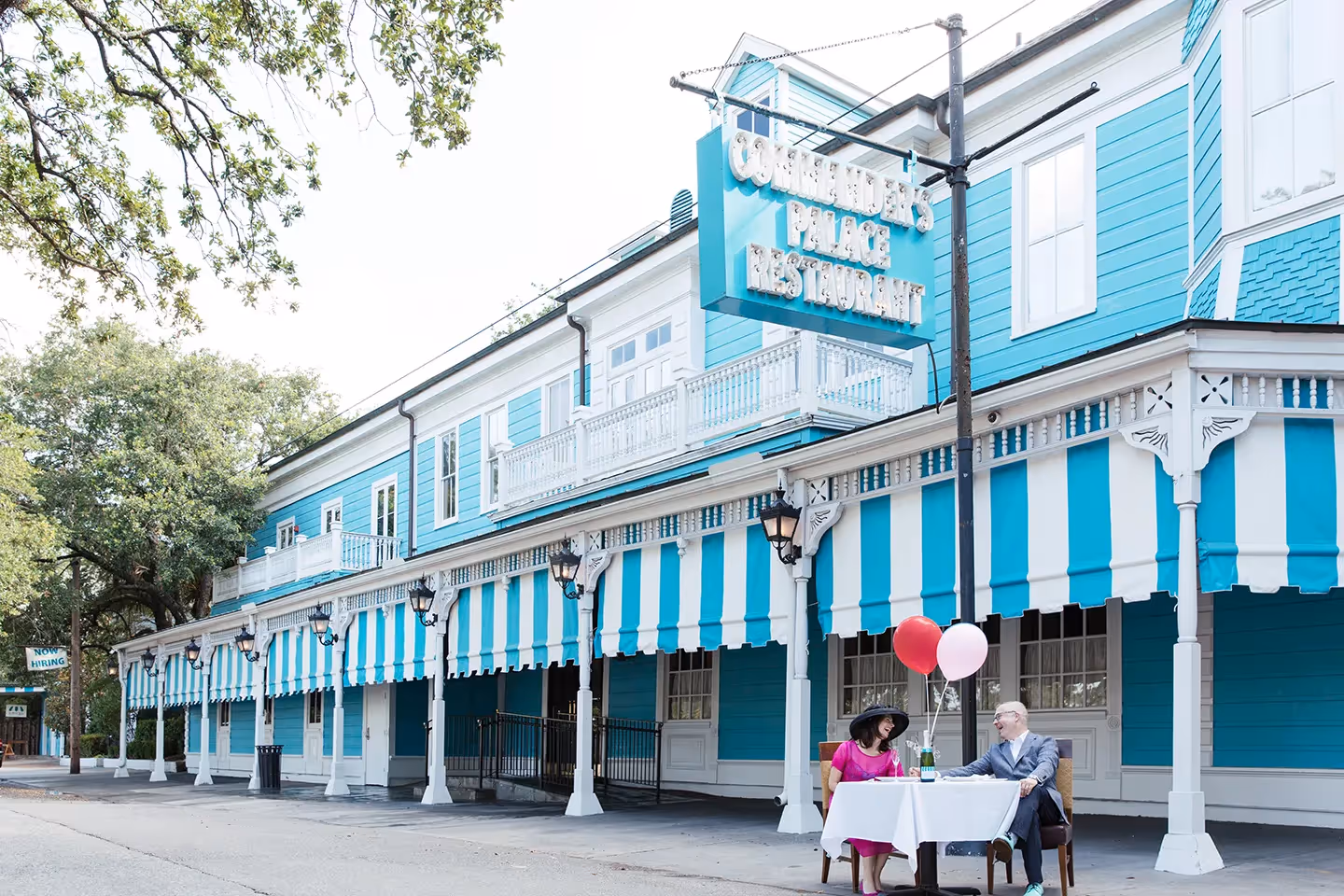 Couple seated at a table outside a blue and white striped awning restaurant named Commander's Palace, with red and pink balloons on the table.