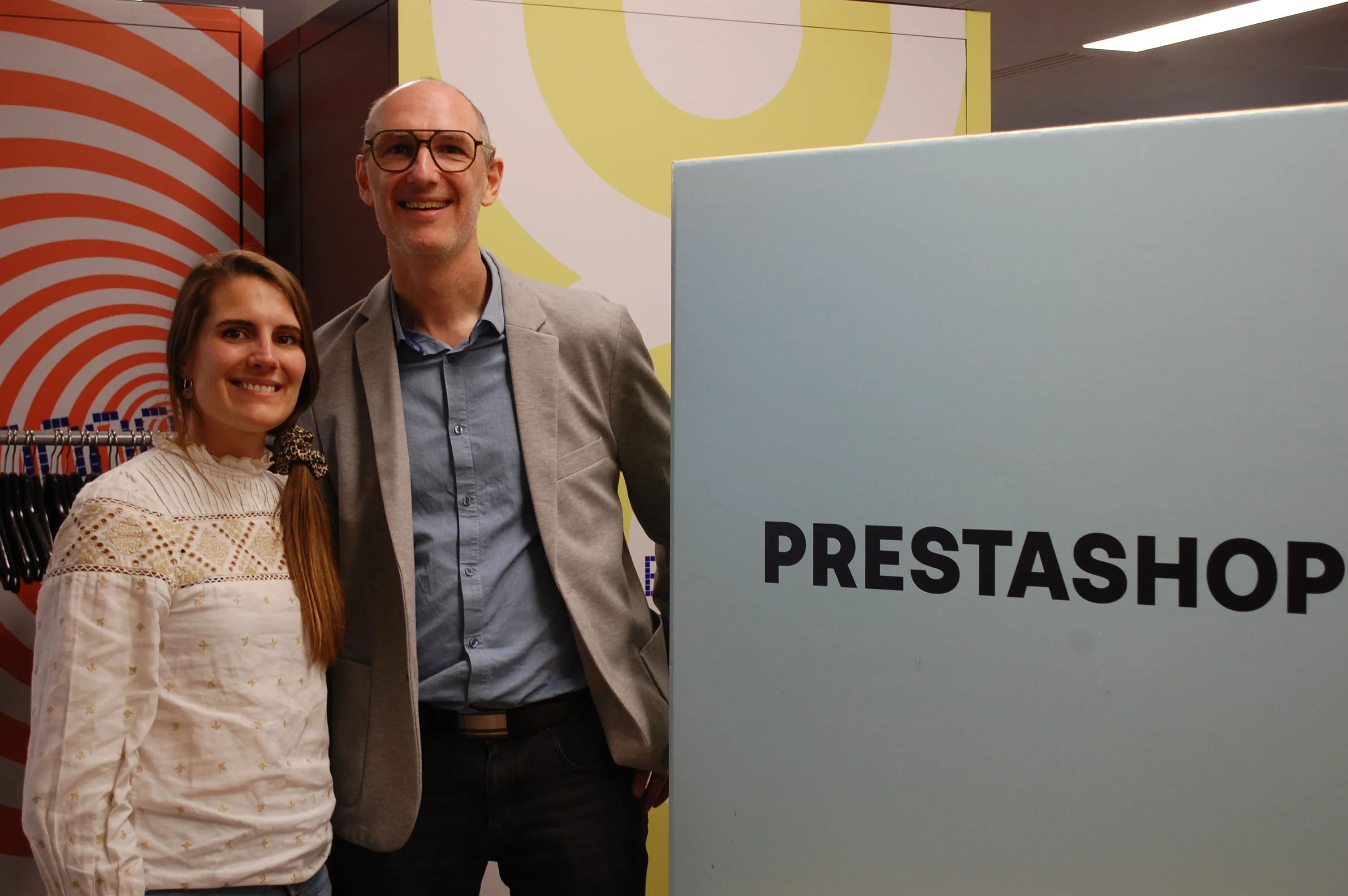 Deux personnes souriantes posent à côté d'un panneau avec le mot PRESTASHOP en noir.