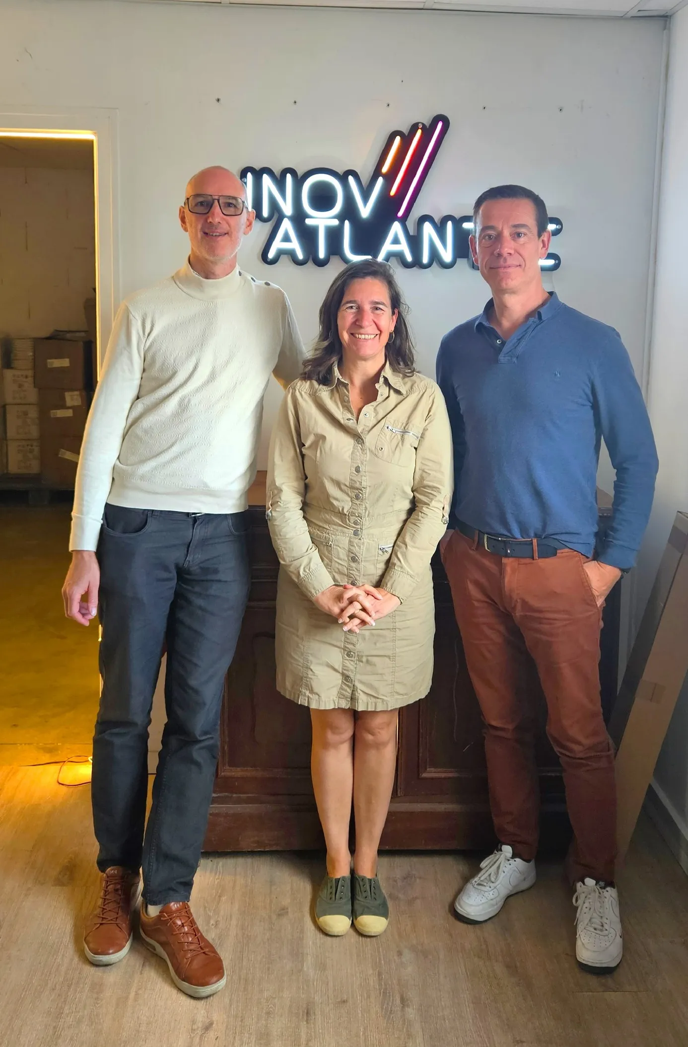 Trois personnes debout souriantes devant un mur avec un panneau lumineux INOV ATLANTIC.