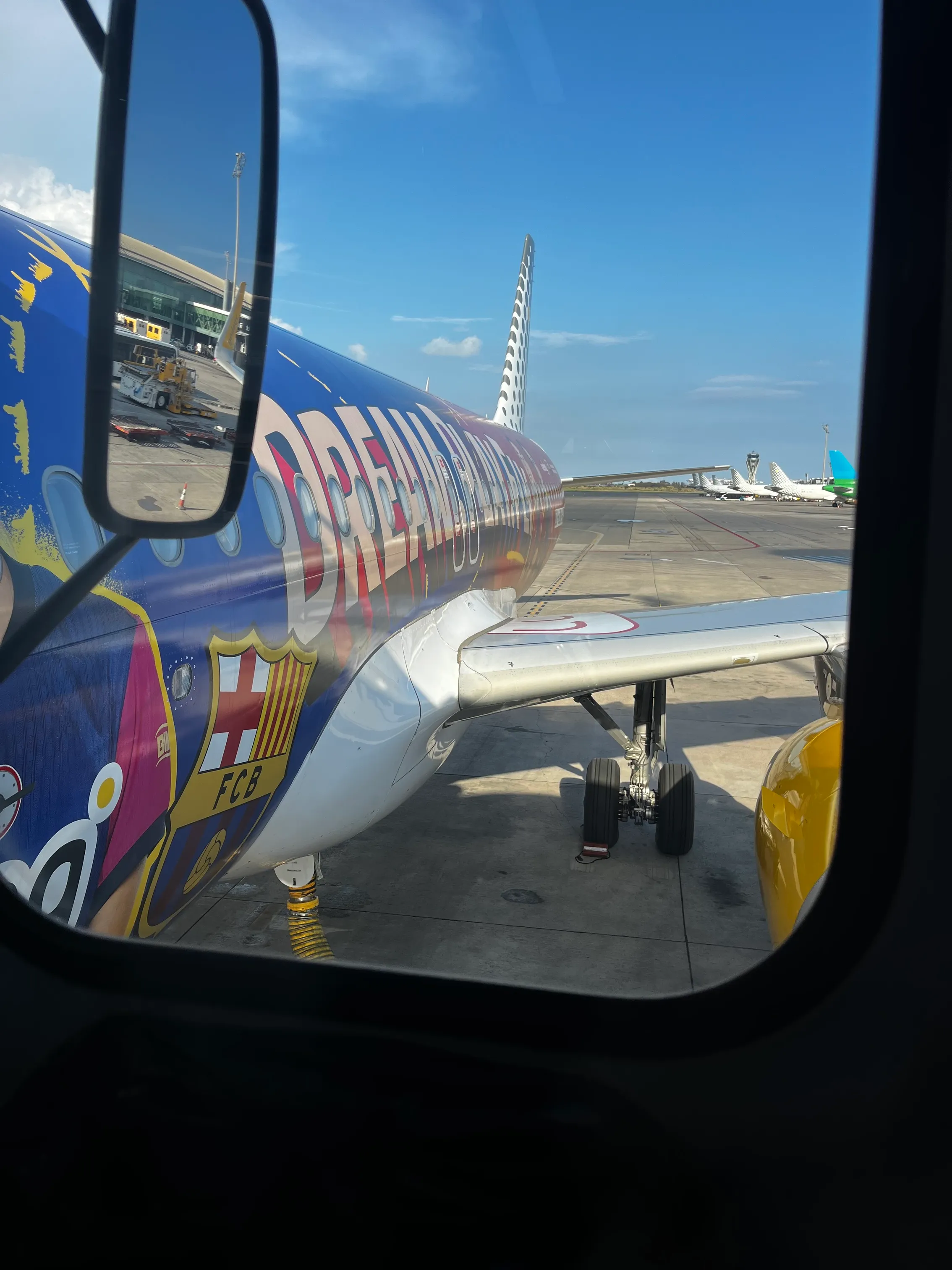 Vue d'un avion coloré Dreamliner avec logo FCB vu d'une fenêtre au sol sous ciel bleu.
