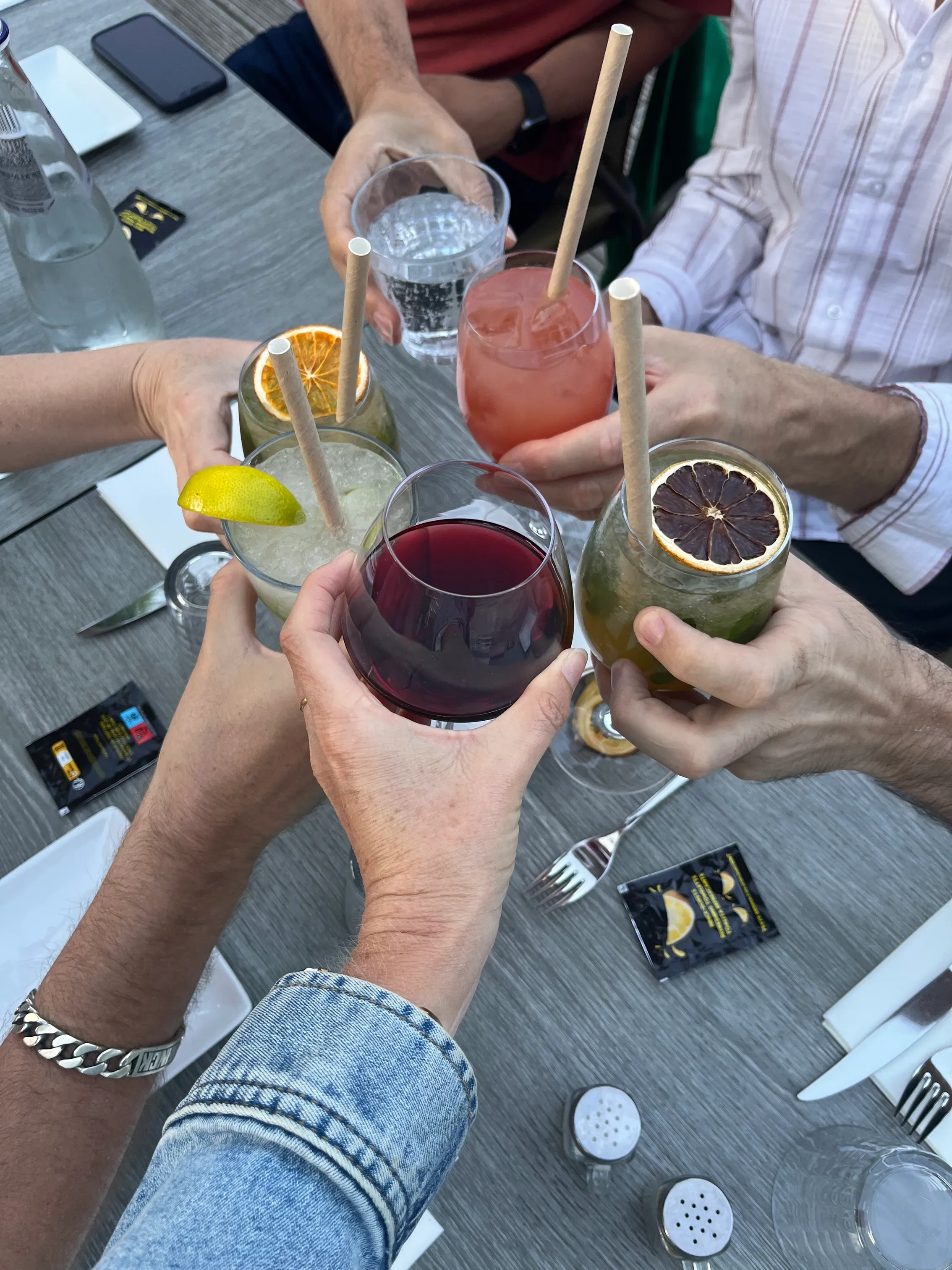 Cinq personnes portent un toast avec des verres de cocktails et un verre de vin rouge au-dessus d'une table.