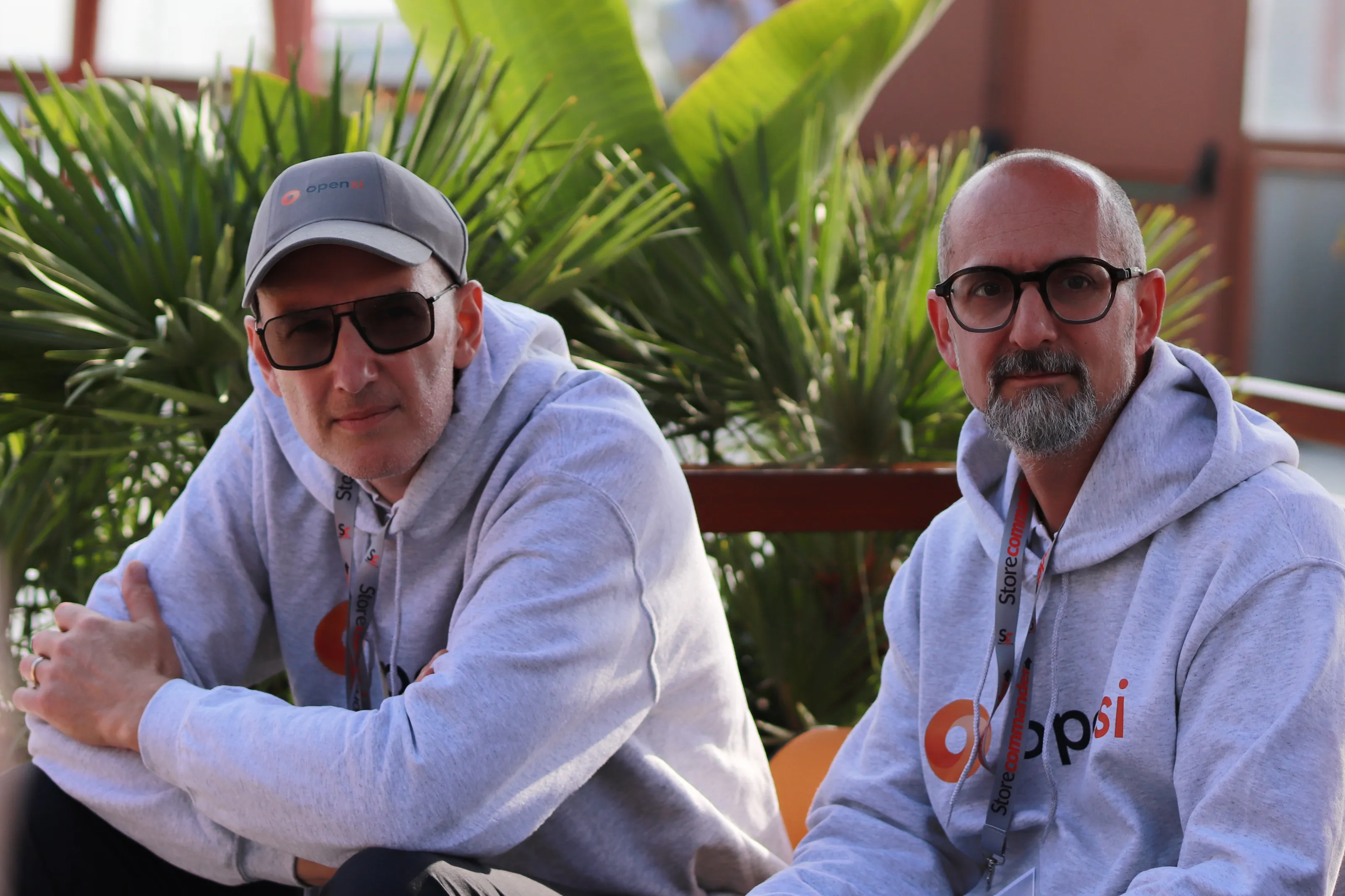 Deux hommes portant des sweat-shirts gris avec logo orange, assis devant des plantes vertes.