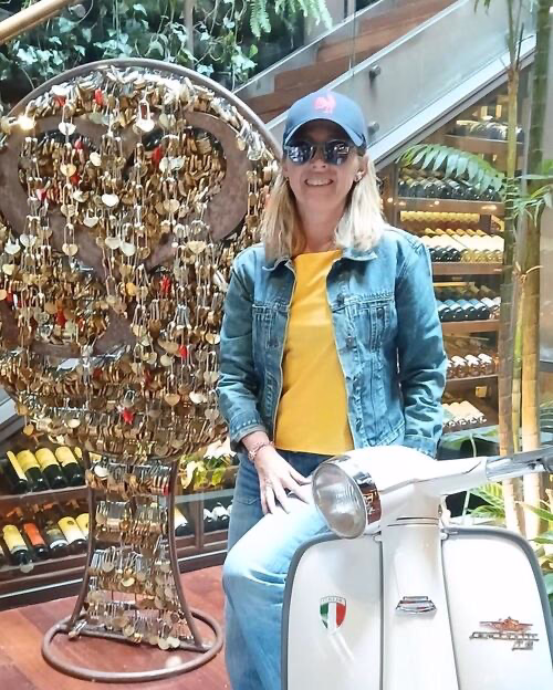 Femme souriante en casquette et veste en jean posant près d'un scooter blanc et d'une sculpture de clés.
