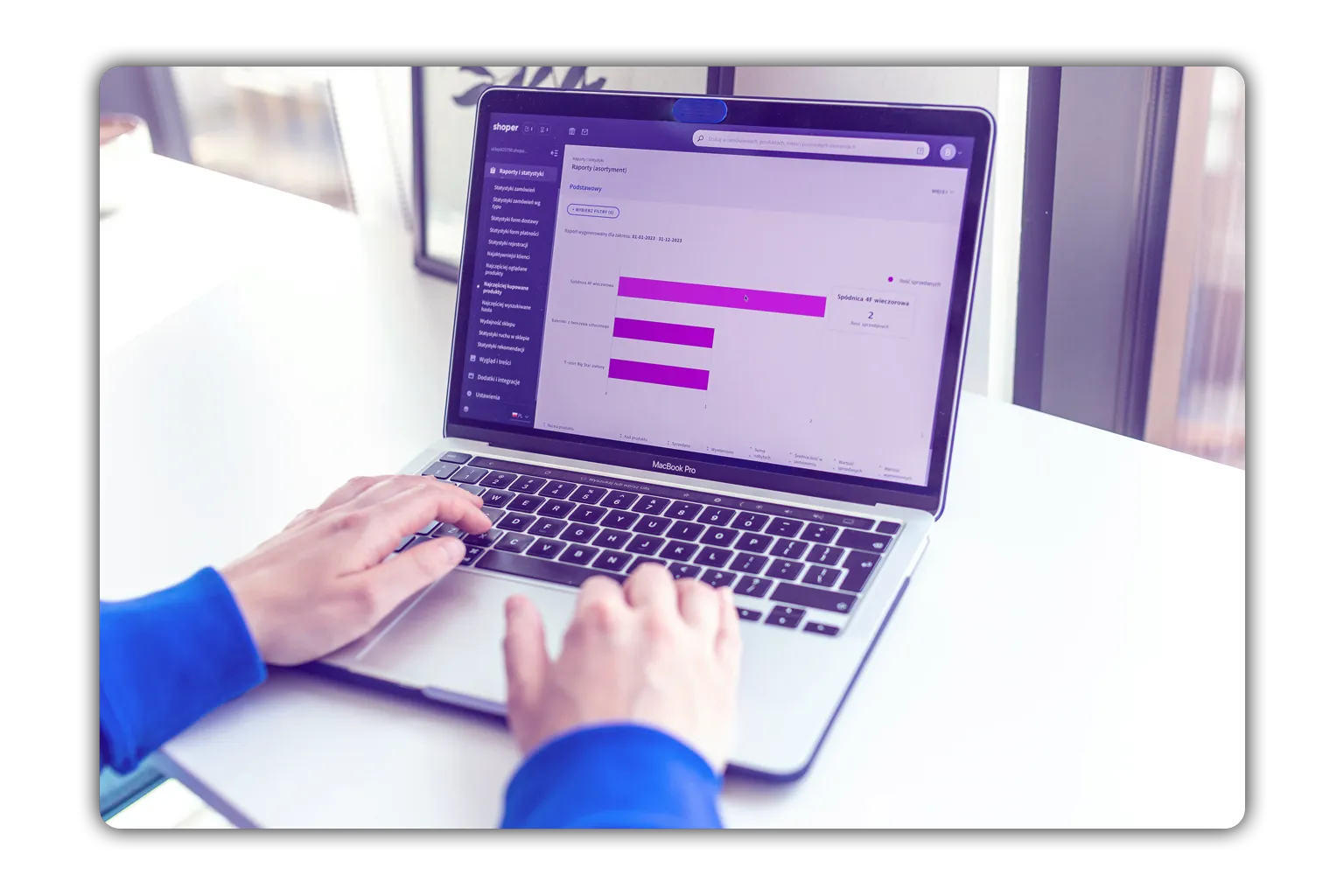 Person typing on a MacBook Pro displaying a sales report with purple bar charts on a white desk near a window.