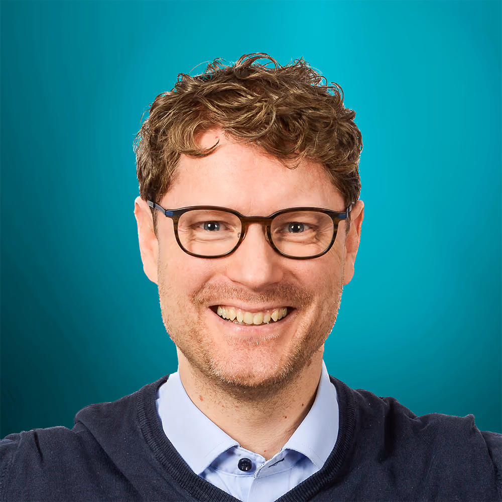 Smilende mann med briller, bølget lyst hår og mørk genser foran blågrønn bakgrunn.