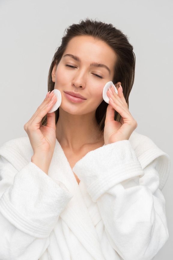 A woman with healthy skin following a skin care routine, gently applying toner to her face with cotton pads. She is wearing a white bathrobe, showcasing her beautiful skin.