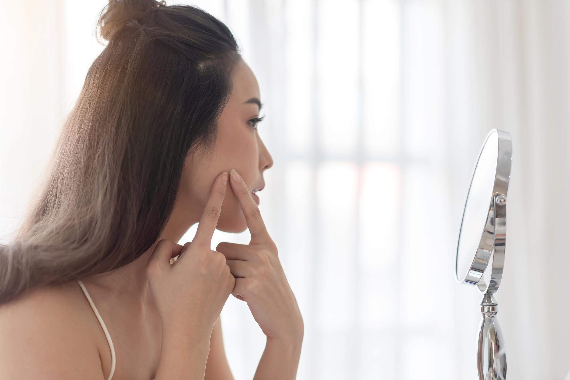 Jeune femme aux longs cheveux bruns scrutant ses pores dilatés sur le visage devant un miroir de salle de bain bien éclairée.