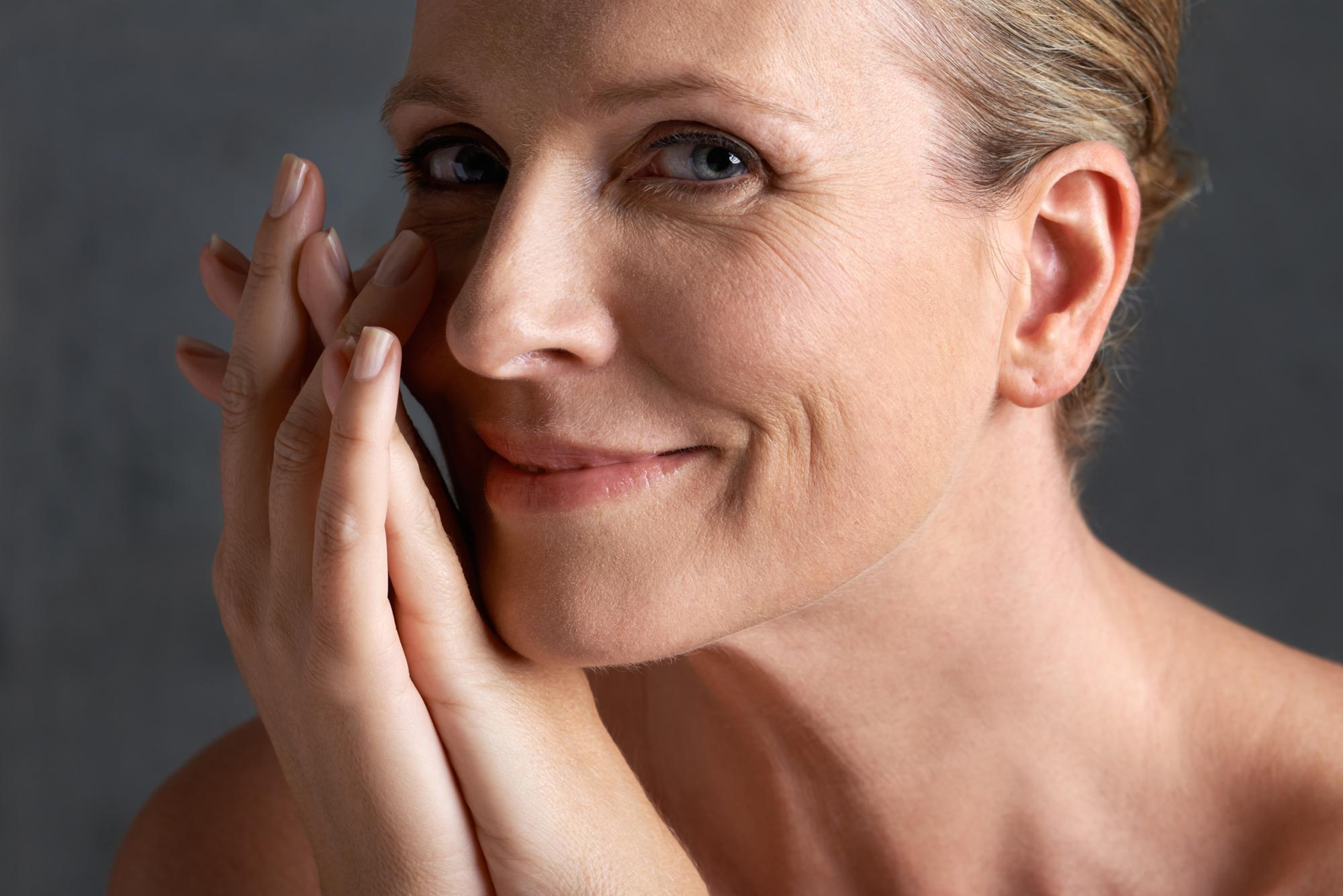 Femme d'âge mûr aux cheveux blonds souriante touchant ses joues avec des rides du visage légères visibles sur fond gris.