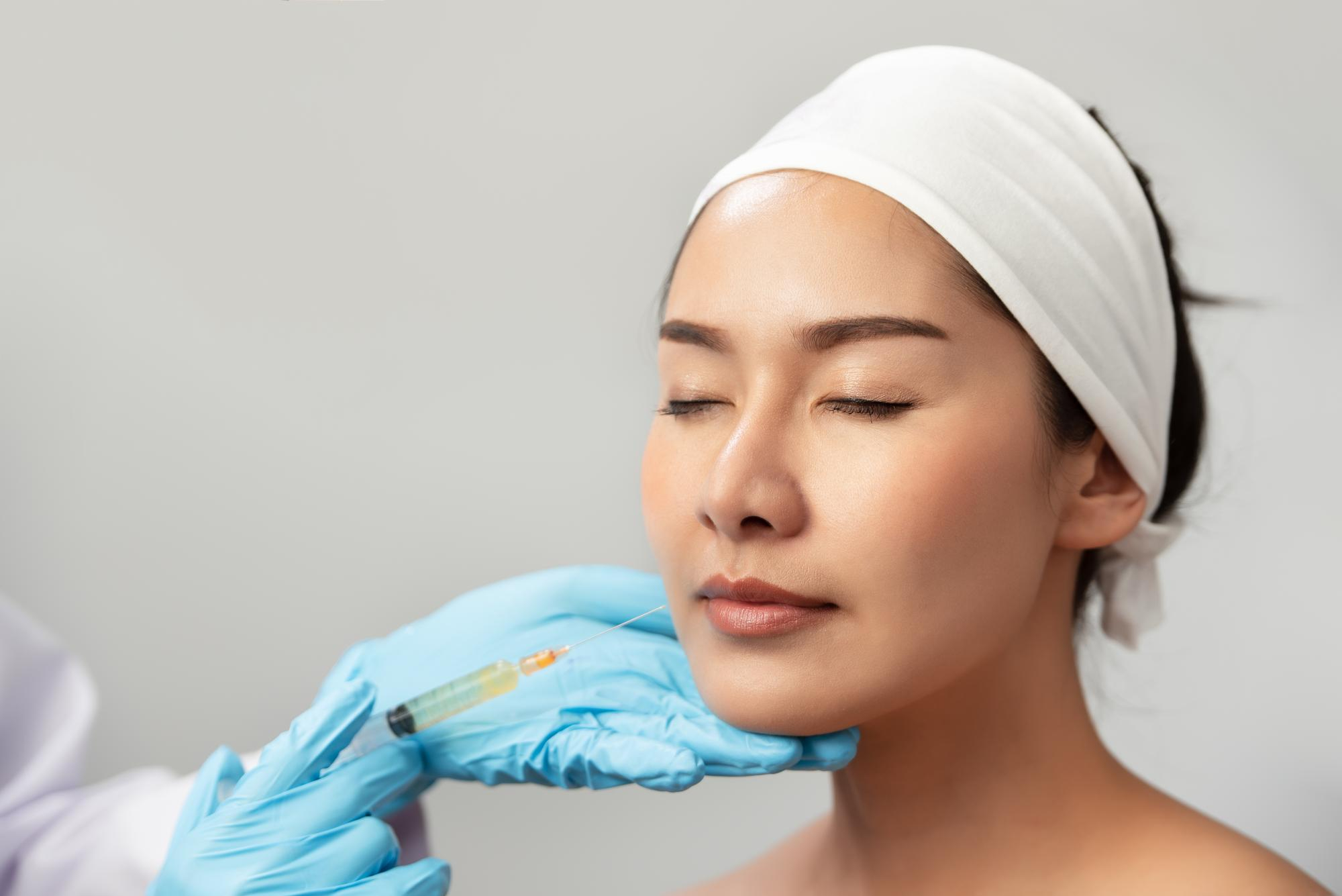 Femme asiatique portant bandeau recevant injection d'agents de comblement ou Botox sur la joue par un professionnel en gants. 