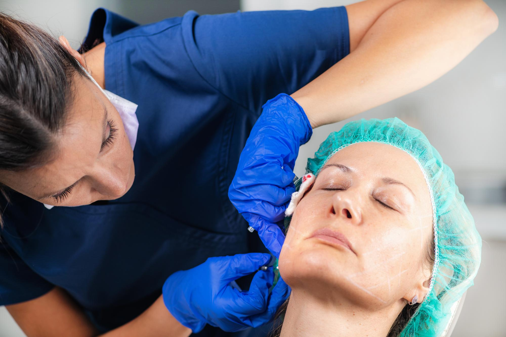 Professionnel médical en tenue bleue et gants effectuant un traitement laser des rides, femme en filet turquoise en clinique.