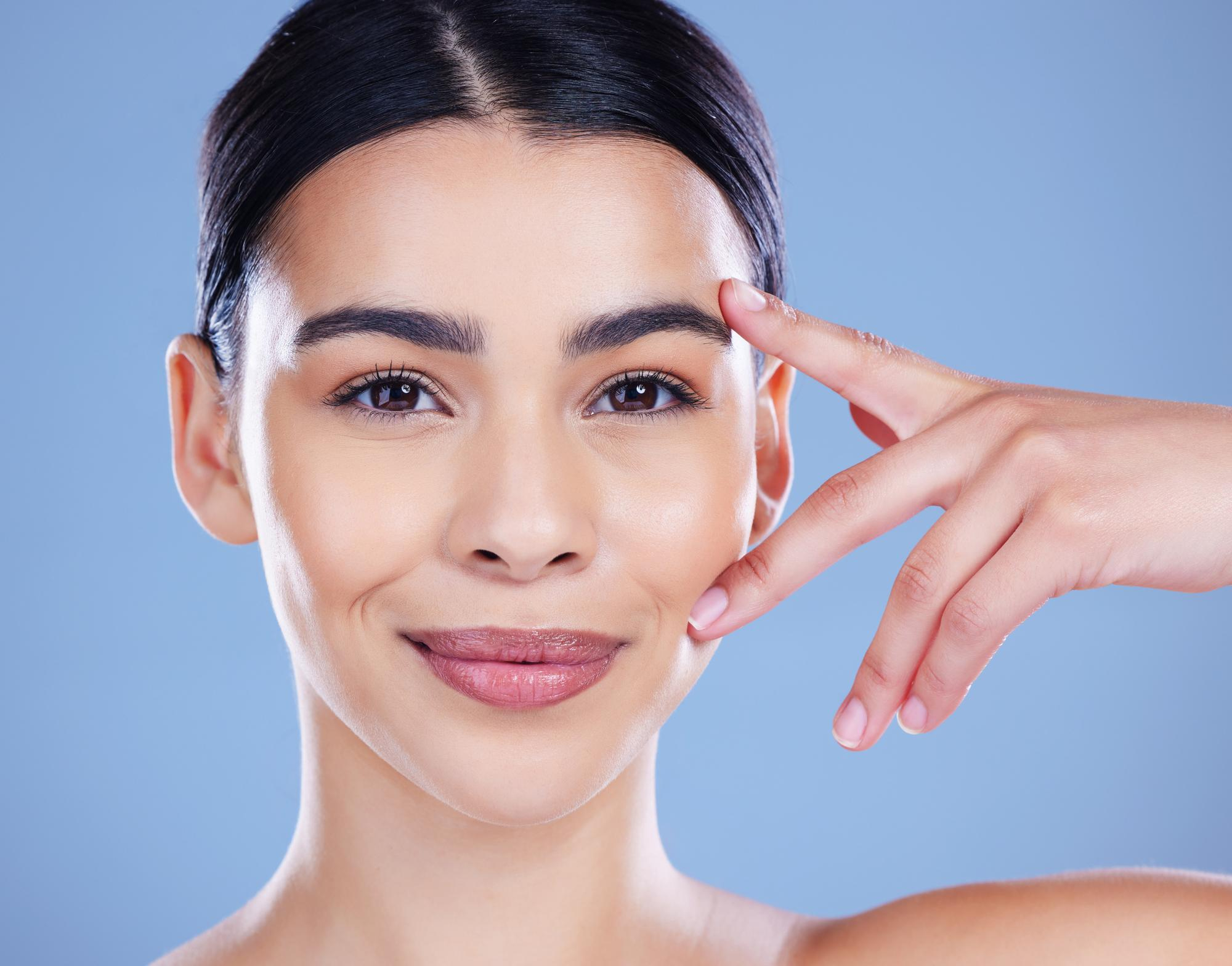 Jeune femme aux cheveux foncés, pointant sa zone oculaire sur fond bleu, montrant les résultats après un traitement des paupières tombantes en clinique.