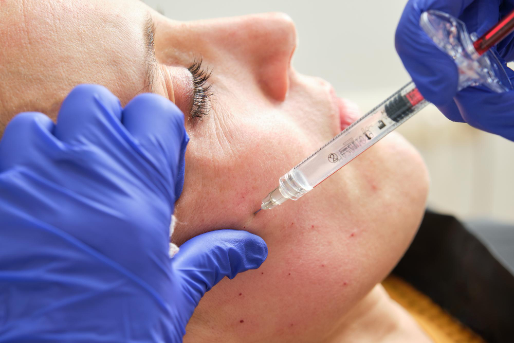 Deux mains gantées de bleu guident une seringue dans la joue au cours d'un traitement par injection au visage, la cliente reste calme.