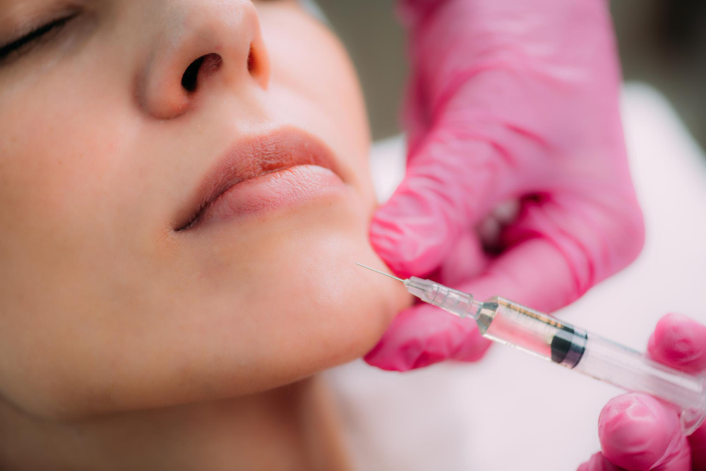 Des mains gantées de rose guident une seringue d'acide hyaluronique dans le menton d'une femme allongée, aux yeux bien fermés.
