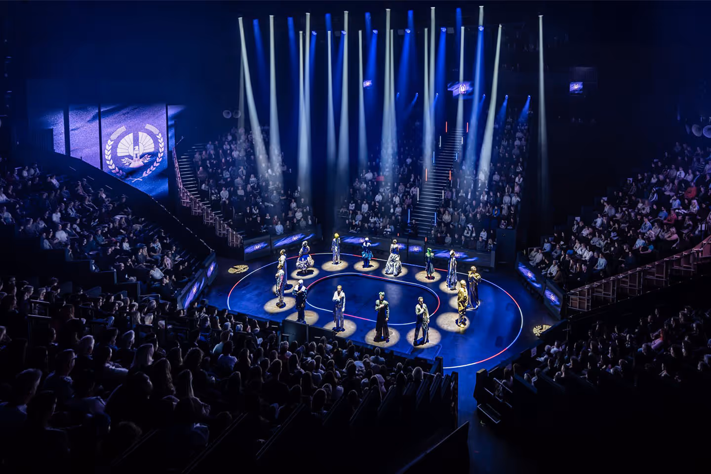 Theatrical stage with performers each standing in individual spotlights surrounded by a seated audience in a darkened auditorium.