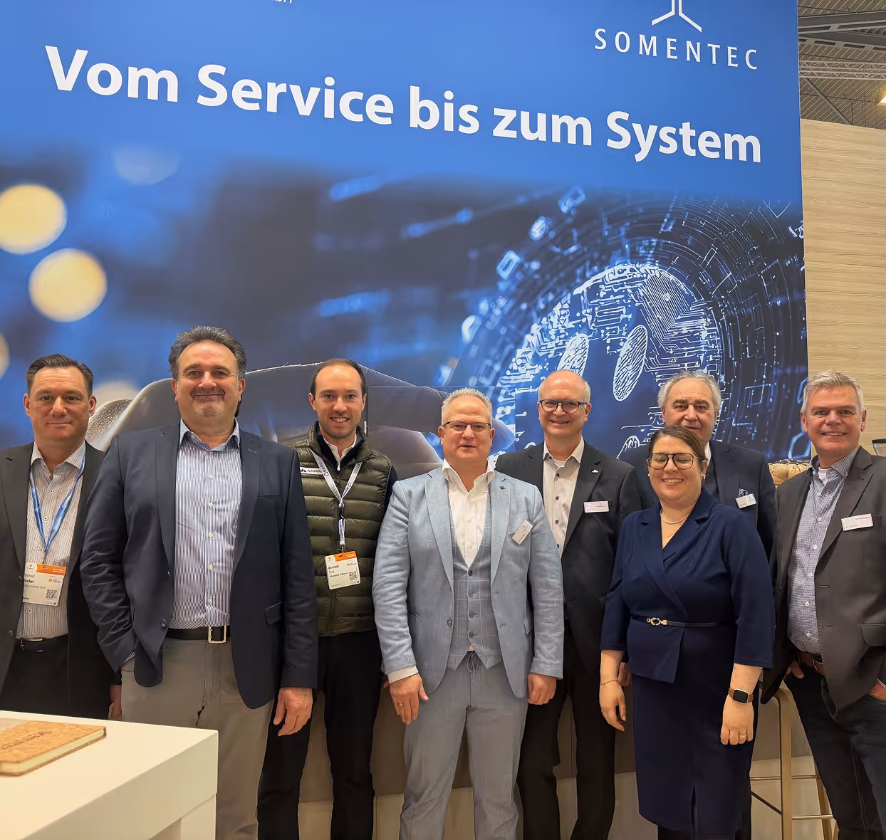 Eight professionals standing in front of a blue SOMENTEC banner that reads 'Vom Service bis zum System,' smiling at the camera at a business event.