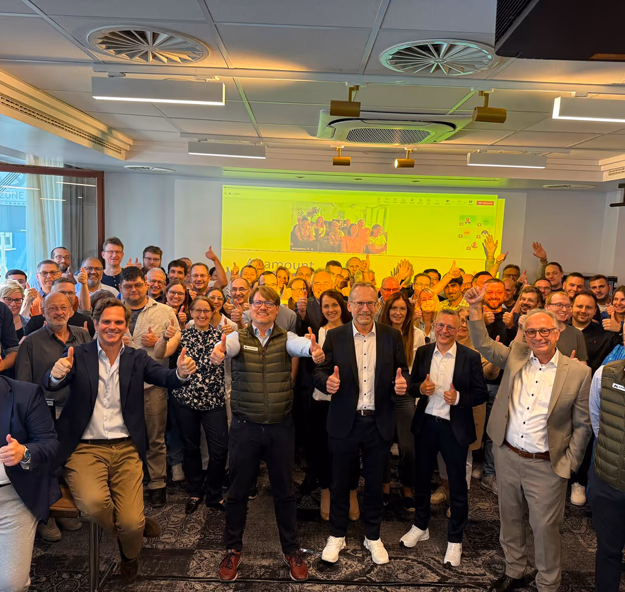 Large group of smiling people indoors giving thumbs up in front of a screen.