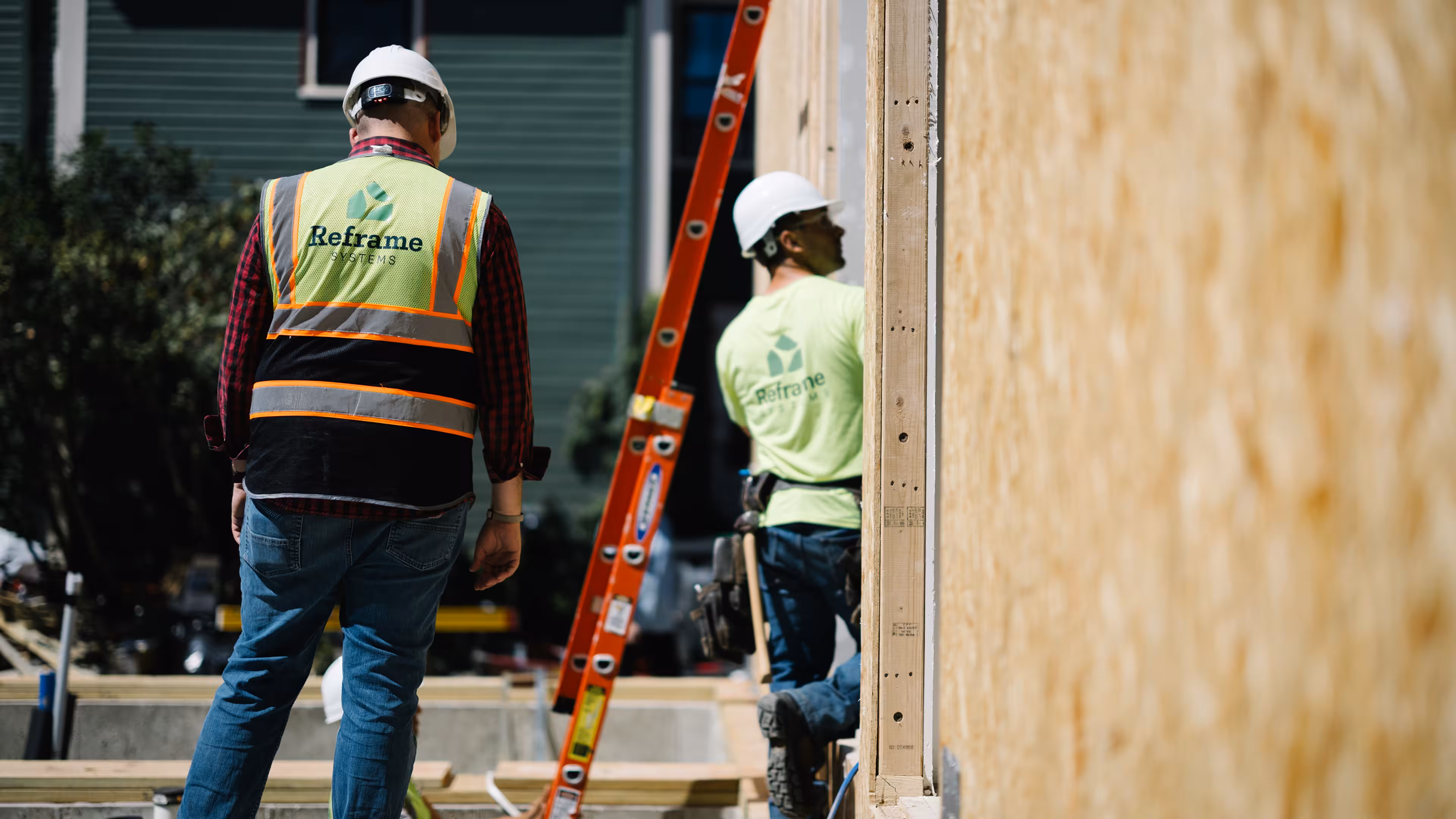 Two Reframe Systems workers in hard hats and safety vests at a wood-framed build site