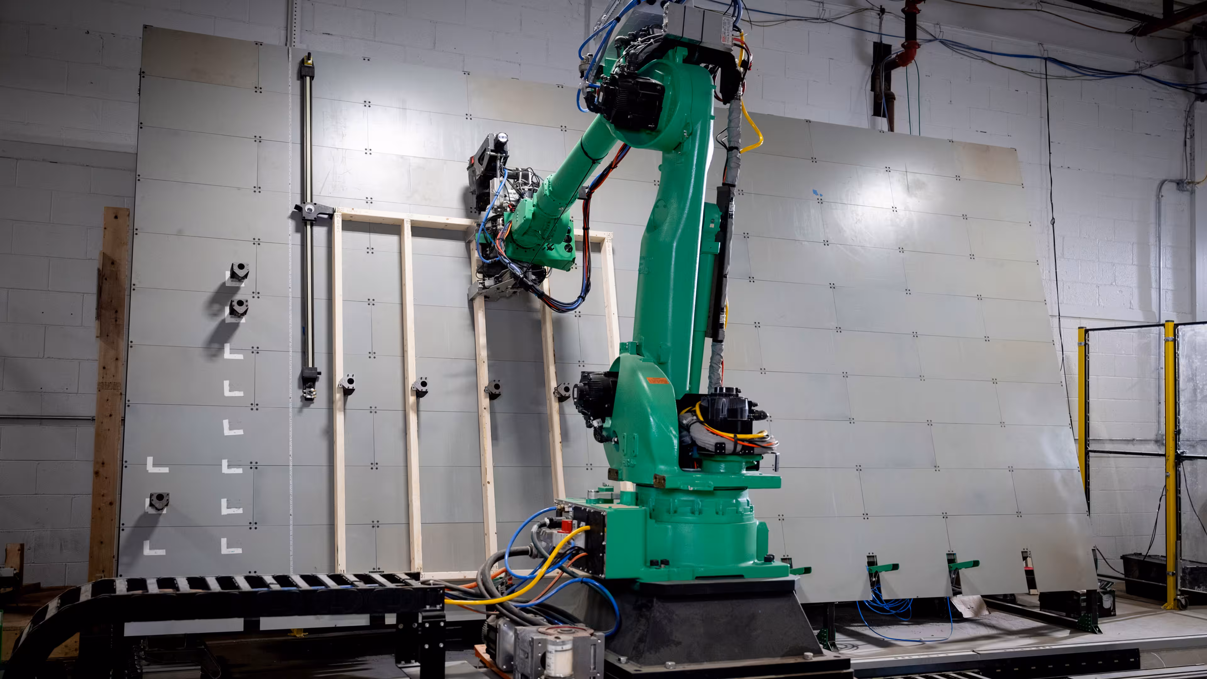 A green industrial robotic arm performing framing work on a wall panel inside a Reframe Systems manufacturing facility.