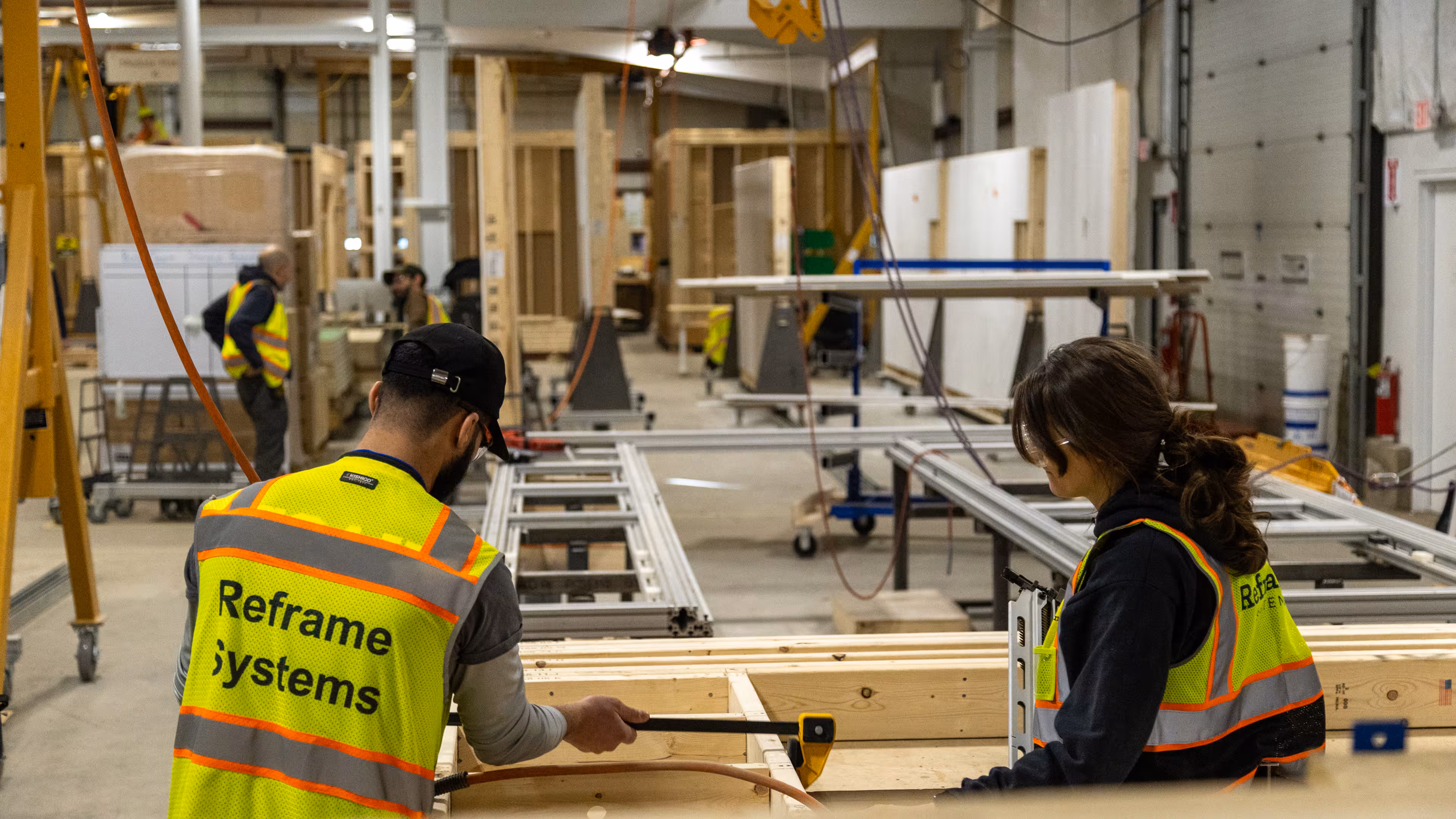 Two Reframe Systems workers in high-visibility vests assembling a wood-framed panel on the factory floor, with additional workers and panel production lines visible in the background.